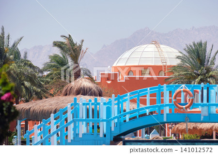 Picture from exotic summer holiday vacations. Pedestrian bridge to beach, building with round roof, palm trees and distant mountains Picture from exotic summer holiday vacations. Pedestrian bridge to beach, building with round roof, palm trees and distant mountains 141012572