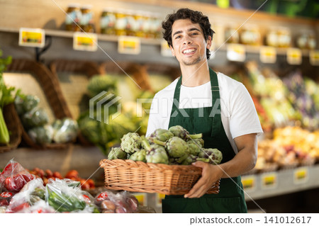 Male seller wearing apron holding artichokes in hand in grocery shop Male seller wearing apron holding artichokes in hand in grocery shop 141012617