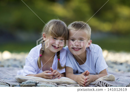 Two young happy cute blond children, boy and girl, brother and sister laying on pebbled beach on blurred bright sunny summer day background. Siblings friendship, leisure and perfect holiday concept. Two young happy cute blond children, boy and girl, brother and sister laying on pebbled beach on blurred bright sunny summer day background. Siblings friendship, leisure and perfect holiday concept. 141012619
