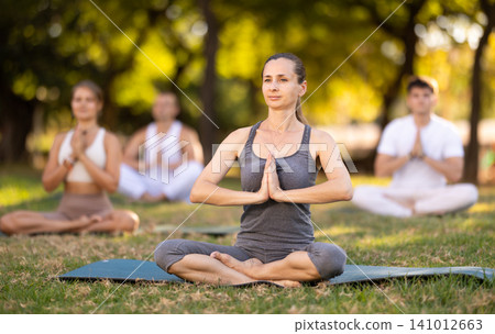 Yoga lesson visitors with girl coach perform padmasana in park Yoga lesson visitors with girl coach perform padmasana in park 141012663