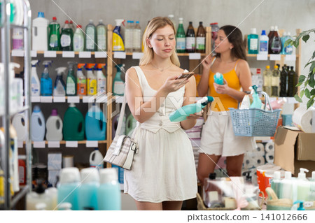 Girl scans QR code on label of window and glass cleaning spray in supermarket. In the background, shoppers are choosing product Girl scans QR code on label of window and glass cleaning spray in supermarket. In the background, shoppers are choosing product 141012666