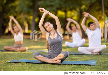 Woman stretching in lotus position during group yoga in park Woman stretching in lotus position during group yoga in park 141012727