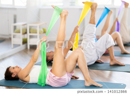 Concentrated active young woman and group of sporty people lying on mats and performing set of pilates exercises with resistance bands in fitness studio 141012866