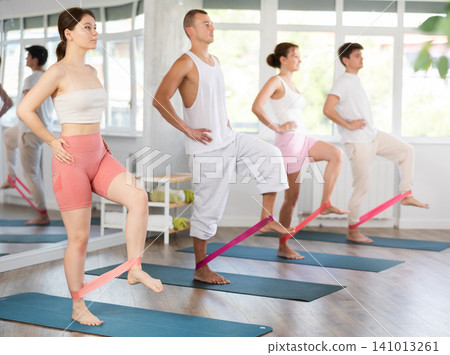Young men and women practicing hip workout with band standing on mat Young men and women practicing hip workout with band standing on mat 141013261