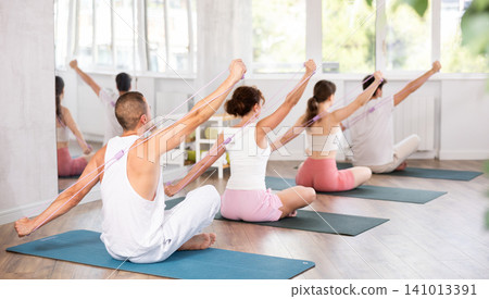 Young men and women doing shoulder workout with resistance band sitting cross-legged 141013391