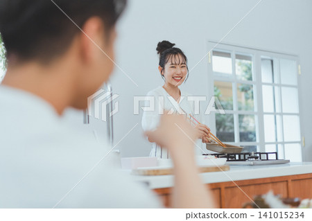 A couple scene in the kitchen: the girlfriend, holding an egg pan and chopsticks, talks to her boyfriend with a big smile on her face. A couple scene in the kitchen: the girlfriend, holding an egg pan and chopsticks, talks to her boyfriend with a big smile on her face. 141015234