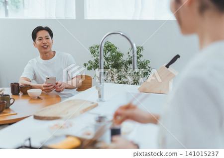A scene of a couple: The boyfriend is smiling and talking to his girlfriend who is cooking in the kitchen while he holds his smartphone in the dining room. A scene of a couple: The boyfriend is smiling and talking to his girlfriend who is cooking in the kitchen while he holds his smartphone in the dining room. 141015237