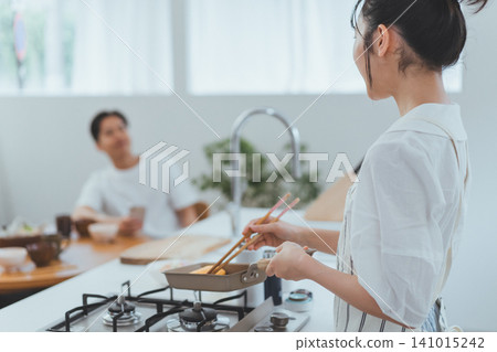 A scene of a couple: a woman turning to face her boyfriend in the kitchen while cooking an omelet, showing her profile, while the boyfriend sits in the dining room. A scene of a couple: a woman turning to face her boyfriend in the kitchen while cooking an omelet, showing her profile, while the boyfriend sits in the dining room. 141015242