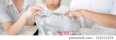 A family of three, including a parent and child, discusses disaster preparedness while checking the emergency supplies in their emergency backpack, without revealing their faces. A family of three, including a parent and child, discusses disaster preparedness while checking the emergency supplies in their emergency backpack, without revealing their faces. 141015259