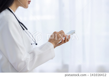 A close-up of a female doctor's hands as she operates her smartphone by the window. A close-up of a female doctor's hands as she operates her smartphone by the window. 141016412