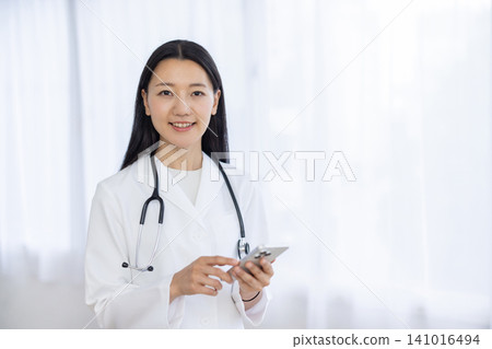 A young Japanese female doctor smiles at the camera while operating her smartphone by the window. 141016494