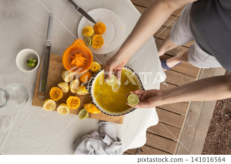 Citrus juicing squeezing limes and oranges into a bowl, in outdoor table setting with wooden deck, with limes, oranges, juicer, bowl of juice, with wood plastic ceramic texture. Citrus juicing squeezing limes and oranges into a bowl, in outdoor table setting with wooden deck, with limes, oranges, juicer, bowl of juice, with wood plastic ceramic texture. 141016564