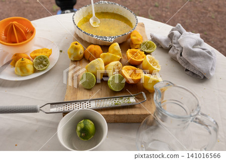Preparing citrus juice squeezing and grating citrus fruits, in outdoor table setup, with juicer, cut lemons, grater, limes, with wooden cutting board, ceramic bowl, glass pitcher texture. 141016566