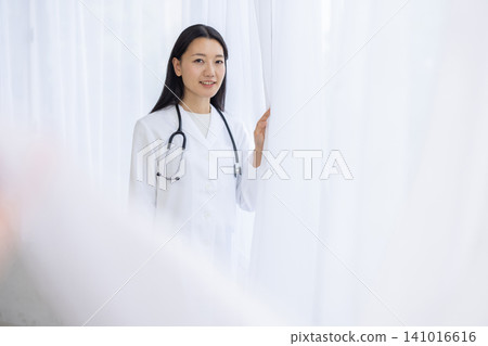 A young Japanese female doctor smiles at the camera, her hand on the curtain, bathed in the light from the window. A young Japanese female doctor smiles at the camera, her hand on the curtain, bathed in the light from the window. 141016616