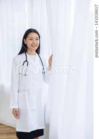 A young Japanese female doctor smiles at the camera, her hand on the curtain, bathed in the light from the window. A young Japanese female doctor smiles at the camera, her hand on the curtain, bathed in the light from the window. 141016617
