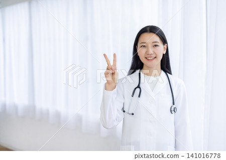 A young Japanese female doctor smiles at the camera, holding up two fingers, in a bright window. A young Japanese female doctor smiles at the camera, holding up two fingers, in a bright window. 141016778