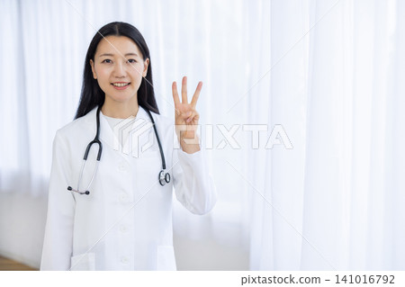 A young Japanese female doctor smiles at the camera, holding up three fingers, in a bright window. A young Japanese female doctor smiles at the camera, holding up three fingers, in a bright window. 141016792