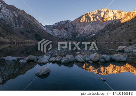 Tranquil mountain lake reflection of golden sunrise over majestic Sierra Nevada. Calm water and rocky shore create peaceful and beautiful outdoor landscape scene Tranquil mountain lake reflection of golden sunrise over majestic Sierra Nevada. Calm water and rocky shore create peaceful and beautiful outdoor landscape scene 141016818