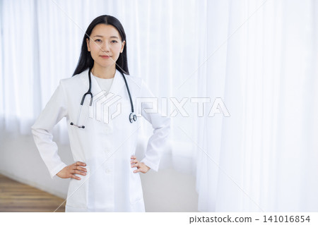 一位年輕的日本女醫生在燈光明亮的窗邊，雙手叉腰，對著鏡頭微笑。 141016854