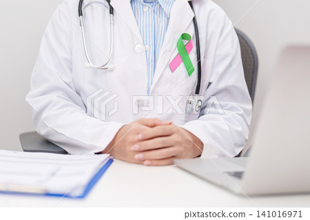 doctor using laptop and report notes on notebook. green and pink ribbon to patient for concept of October Metastatic Breast Cancer Awareness month, National Cancer Survivors Month and Pink October doctor using laptop and report notes on notebook. green and pink ribbon to patient for concept of October Metastatic Breast Cancer Awareness month, National Cancer Survivors Month and Pink October 141016971