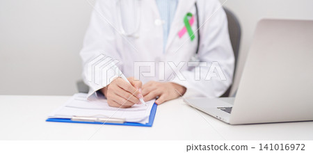 doctor using laptop and report notes on notebook. green and pink ribbon to patient for concept of October Metastatic Breast Cancer Awareness month, National Cancer Survivors Month and Pink October doctor using laptop and report notes on notebook. green and pink ribbon to patient for concept of October Metastatic Breast Cancer Awareness month, National Cancer Survivors Month and Pink October 141016972