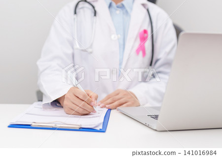 Professional doctor wearing lab coat using laptop and taking report notes on notebook for Breast Cancer Awareness month, World Cancer Day, National Cancer Survivors Month and Pink October in hospital Professional doctor wearing lab coat using laptop and taking report notes on notebook for Breast Cancer Awareness month, World Cancer Day, National Cancer Survivors Month and Pink October in hospital 141016984