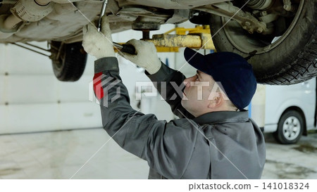 Car mechanic wearing grey uniform repairing vehicle, working underneath lifted car and adjusting bolts with wrench in automotive workshop 141018324