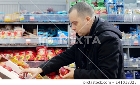 Man picking ripe red apples from colorful produce section, surrounded by packaged groceries, focusing on nutritious selection in modern supermarket Man picking ripe red apples from colorful produce section, surrounded by packaged groceries, focusing on nutritious selection in modern supermarket 141018325