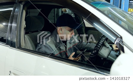 Car mechanic sitting inside vehicle at auto repair shop, using smartphone for accessing repair details or communicating with work team Car mechanic sitting inside vehicle at auto repair shop, using smartphone for accessing repair details or communicating with work team 141018327