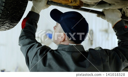 Professional mechanic wearing gloves and baseball cap is working under a lifted car, holding a metal bar and repairing the car's bottom in a professional auto repair shop 141018328