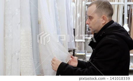 Male shopper carefully feeling white curtain fabric, evaluating texture and quality in textile store, considering home decor and interior design selection Male shopper carefully feeling white curtain fabric, evaluating texture and quality in textile store, considering home decor and interior design selection 141018339