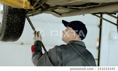 Car mechanic in grey uniform and blue cap leveraging crowbar while repairing vehicle inside clean, modern automotive workshop Car mechanic in grey uniform and blue cap leveraging crowbar while repairing vehicle inside clean, modern automotive workshop 141018345
