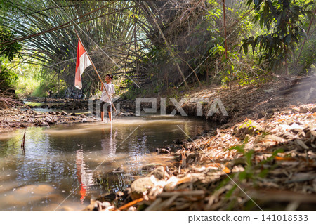 Young person standing in shallow forest stream holding red and white flag beneath dense tropical branches, with sunlight reflecting on water and calm remote wilderness atmosphere 141018533