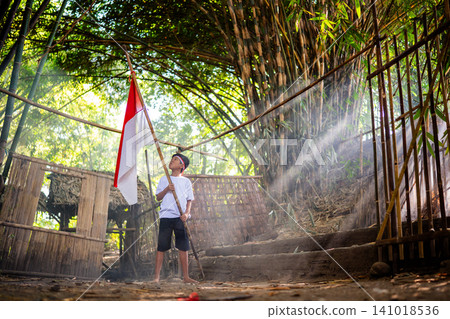 Young boy standing with red and white flag inside rustic bamboo shelter with sunlight beams, dusty ground, steps, and natural village surroundings in peaceful outdoor scene Young boy standing with red and white flag inside rustic bamboo shelter with sunlight beams, dusty ground, steps, and natural village surroundings in peaceful outdoor scene 141018536