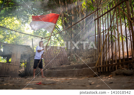 Young boy holding red and white flag inside bamboo village courtyard with sunlight streaming through trees, rustic huts, smoky morning atmosphere, barefoot stance, rural celebration mood Young boy holding red and white flag inside bamboo village courtyard with sunlight streaming through trees, rustic huts, smoky morning atmosphere, barefoot stance, rural celebration mood 141018542