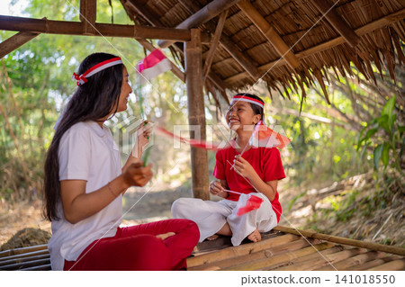 Smiling mother and daughter playing with small red and white flags inside rustic bamboo shelter, sharing joyful family time in warm sunlight with natural outdoor background Smiling mother and daughter playing with small red and white flags inside rustic bamboo shelter, sharing joyful family time in warm sunlight with natural outdoor background 141018550