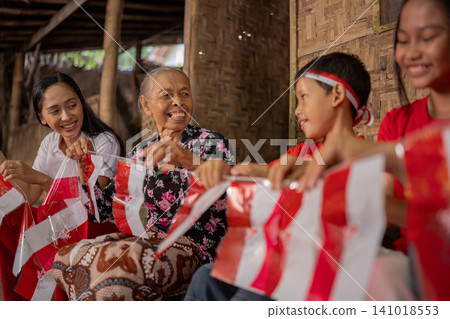 Smiling multigenerational family sharing red and white flags during community celebration inside rustic bamboo home, with grandmother, children and young woman gathered together Smiling multigenerational family sharing red and white flags during community celebration inside rustic bamboo home, with grandmother, children and young woman gathered together 141018553