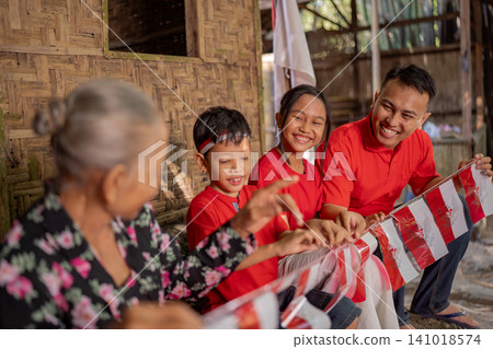 Smiling family in red shirts shares festive moment beside red and white bunting inside rustic bamboo home, with grandmother in foreground and warm natural light creating joyful community atmosphere Smiling family in red shirts shares festive moment beside red and white bunting inside rustic bamboo home, with grandmother in foreground and warm natural light creating joyful community atmosphere 141018574