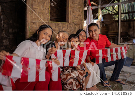 Smiling multigenerational family holding red and white flags beside rustic wooden home during warm daylight, festive outdoor gathering with children, elders, casual clothing, rural community spirit Smiling multigenerational family holding red and white flags beside rustic wooden home during warm daylight, festive outdoor gathering with children, elders, casual clothing, rural community spirit 141018586
