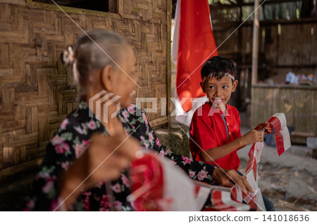Smiling young boy in red shirt holds red and white flags while seated beside older woman in rustic bamboo home, with warm daylight and festive village atmosphere Smiling young boy in red shirt holds red and white flags while seated beside older woman in rustic bamboo home, with warm daylight and festive village atmosphere 141018636
