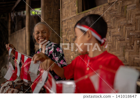 Smiling elderly woman sharing festive red and white bunting with child beside rustic bamboo wall in warm village home, capturing family celebration, cultural tradition, and joyful community moment 141018647