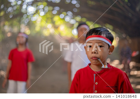 Young boy in red shirt and headband holds spoon in mouth while standing with blurred children outdoors under dappled light on shaded path Young boy in red shirt and headband holds spoon in mouth while standing with blurred children outdoors under dappled light on shaded path 141018648