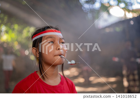 Young girl in red shirt and headband eating from spoon during outdoor gathering in soft sunlight, with blurred background, candid expression, playful childhood mood, close portrait composition Young girl in red shirt and headband eating from spoon during outdoor gathering in soft sunlight, with blurred background, candid expression, playful childhood mood, close portrait composition 141018652