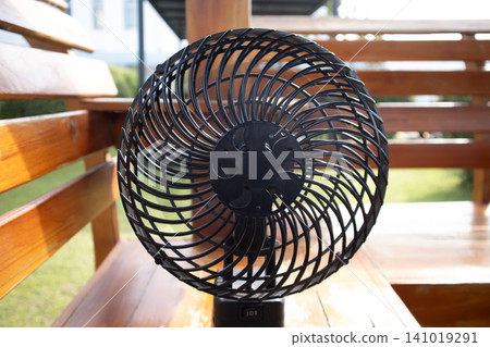 Black electric fan spiral grille wooden bench outdoor seating summer cooling portable appliance metal frame front view sunlight Black electric fan spiral grille wooden bench outdoor seating summer cooling portable appliance metal frame front view sunlight 141019291