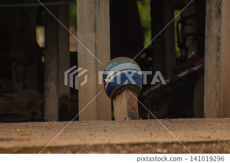 Blue helmet on wooden post in rustic shelter, worn protective gear in dark rural setting with sandy ground and weathered structure Blue helmet on wooden post in rustic shelter, worn protective gear in dark rural setting with sandy ground and weathered structure 141019296