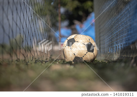 Soccer ball on grass near net in outdoor field with blurred background and shallow depth of field, sports equipment resting after play in worn condition Soccer ball on grass near net in outdoor field with blurred background and shallow depth of field, sports equipment resting after play in worn condition 141019311
