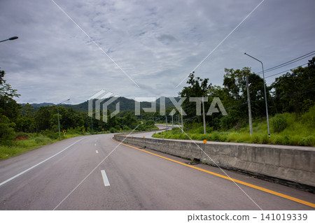 Curved empty road through green forest under cloudy sky Curved empty road through green forest under cloudy sky 141019339
