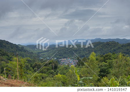 Mountain valley village under cloudy sky, lush green forest, distant ridge, tropical landscape, rural settlement 141019347