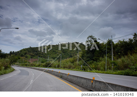 Winding road through green hillside under cloudy sky with empty lane and roadside pole in rural landscape Winding road through green hillside under cloudy sky with empty lane and roadside pole in rural landscape 141019348