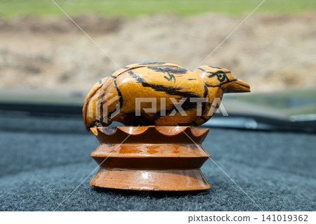 Wooden bird figurine on carved pedestal with blurred outdoor background Wooden bird figurine on carved pedestal with blurred outdoor background 141019362